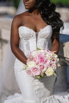 A bride in a white, intricately designed strapless wedding dress holds a bouquet of pink and white roses. Her long, dark hair is styled in loose curls, and the scene appears outdoors with soft, natural lighting.