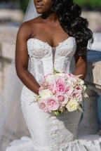 A bride in a white, intricately designed strapless wedding dress holds a bouquet of pink and white roses. Her long, dark hair is styled in loose curls, and the scene appears outdoors with soft, natural lighting.