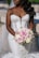 A bride in a white, intricately designed strapless wedding dress holds a bouquet of pink and white roses. Her long, dark hair is styled in loose curls, and the scene appears outdoors with soft, natural lighting.