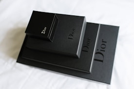 A stack of black boxes of varying sizes, each prominently featuring the Dior logo in elegant lettering. The boxes are arranged on a light-colored surface, creating a contrast with their dark, matte finish.