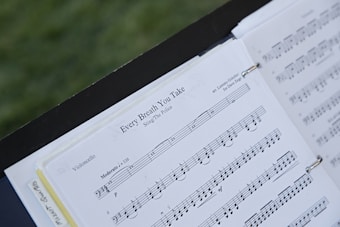A sheet of music is open on a stand outdoors, featuring the piece 'Every Breath You Take' arranged for violoncello by Lorenzo Golofeev. The pages are held in place with rings, and the background is a blurred green, likely grass.