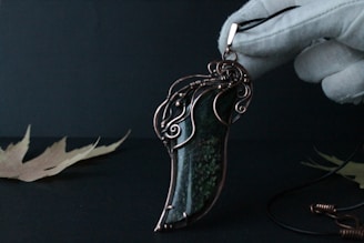 An intricately designed pendant with a polished stone centerpiece, wrapped in ornate metalwork, resembling swirls and curls. The setting is minimalist, with a pair of dried leaves on a dark background. A gloved hand holds the pendant, enhancing its delicate appearance.