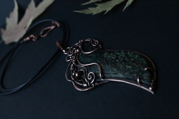 A dark green pendant with intricate wire wrapping rests on a black cord, set against a dark background with subtle leaf patterns.