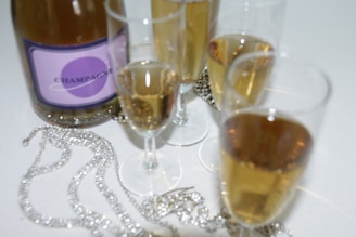 A bottle of champagne with a purple label is surrounded by several filled champagne flutes. Sparkling jewelry pieces are elegantly placed on the white surface next to the glasses, adding a touch of luxury.