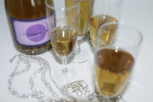 A bottle of champagne with a purple label is surrounded by several filled champagne flutes. Sparkling jewelry pieces are elegantly placed on the white surface next to the glasses, adding a touch of luxury.
