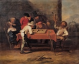 A group of seven people are gathered around a table with an orange tablecloth, intensely observing and playing a board game. One person in a white outfit appears to be making a move, while others watch with interest. One man seated to the right is holding up a mug, seemingly in a celebratory or triumphant gesture. There are various expressions of concentration and engagement among the players. The background includes rustic and earthy tones, suggesting an indoor setting with a large support beam and a scenic view of mountains in the distance.