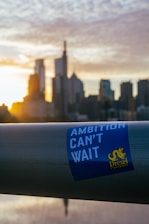 A metal railing with a sticker that reads 'Ambition Can't Wait' with the Drexel University logo is in the foreground. In the background, a city skyline is visible with tall buildings silhouetted against a setting or rising sun, casting a warm glow.