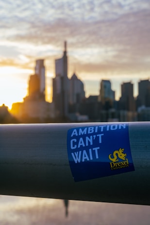 A metal railing with a sticker that reads 'Ambition Can't Wait' with the Drexel University logo is in the foreground. In the background, a city skyline is visible with tall buildings silhouetted against a setting or rising sun, casting a warm glow.