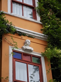 An ornate building facade featuring a prominent orange wall with white trim. A decorative bust is mounted above a window with red, blue, and green panes. Lush green foliage is growing around the structure, adding a natural element to the scene.