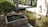A tranquil scene of a small concrete water feature within a garden, with a gently flowing stream and clear reflections. Surrounding the water feature are lush green shrubs and trees, providing a serene environment. Sunlight casts soft shadows on the ground and water, adding a peaceful ambiance.