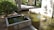 A tranquil scene of a small concrete water feature within a garden, with a gently flowing stream and clear reflections. Surrounding the water feature are lush green shrubs and trees, providing a serene environment. Sunlight casts soft shadows on the ground and water, adding a peaceful ambiance.