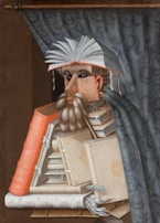 A surreal portrait made from a stack of books, forming the figure of a person with an open book as a hat. The person's face is crafted from book spines and pages, and there is a curtain in the background.