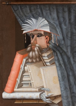 A surreal portrait made from a stack of books, forming the figure of a person with an open book as a hat. The person's face is crafted from book spines and pages, and there is a curtain in the background.