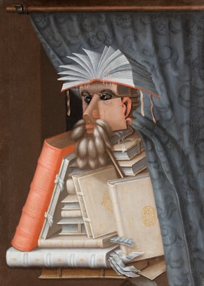 A surreal portrait made from a stack of books, forming the figure of a person with an open book as a hat. The person's face is crafted from book spines and pages, and there is a curtain in the background.