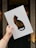 A hand holds a piece of paper with a cutout silhouette of a cat against a blurred, warmly lit background. The paper is white, allowing the colors from the background to be visible through the cat-shaped cutout.