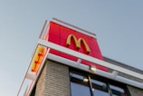 A McDonald's sign with the iconic yellow 'M' logo is prominently displayed on a building's facade. The structure is modern, with a combination of red panels and brick walls, and is taken from a low angle against a clear blue sky.