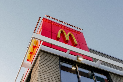A McDonald's sign with the iconic yellow 'M' logo is prominently displayed on a building's facade. The structure is modern, with a combination of red panels and brick walls, and is taken from a low angle against a clear blue sky.