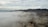 A dense layer of clouds or fog covers the landscape, partially obscuring the view. Mountains rise in the background, with their rugged terrain visible through the haze. Below, some buildings can be seen peeking through the mist, suggesting a semi-industrial area.