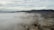 A dense layer of clouds or fog covers the landscape, partially obscuring the view. Mountains rise in the background, with their rugged terrain visible through the haze. Below, some buildings can be seen peeking through the mist, suggesting a semi-industrial area.