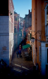A narrow, sunlit street flanked by colorful old buildings with flat roofs and iron decorations. A few people walk down the cobblestone path, which has shadows cast by the buildings. The street is lined with outdoor seating and an awning, enhancing the charming atmosphere.