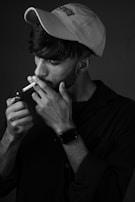A person wearing a cap and a long-sleeved shirt is holding a cigarette close to their mouth and lighting it with a small flame. The monochrome setting highlights the contours and expressions, giving a dramatic feel to the scene.
