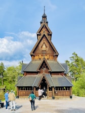 A wooden stave church with intricate details, characterized by its steep gabled roof and dark brown color. The church is surrounded by lush greenery, and several people are casually standing and interacting near the entrance.