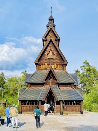 A wooden stave church with intricate details, characterized by its steep gabled roof and dark brown color. The church is surrounded by lush greenery, and several people are casually standing and interacting near the entrance.