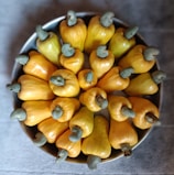 A bowl filled with bright yellow cashew apples, each with a distinctive green-gray cashew nut attached at the top. The cashew apples are arranged in a circular pattern, creating an eye-catching display.