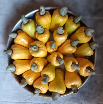 A bowl filled with bright yellow cashew apples, each with a distinctive green-gray cashew nut attached at the top. The cashew apples are arranged in a circular pattern, creating an eye-catching display.