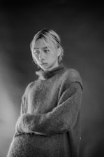 A monochrome portrait of a person with light-colored hair wearing a textured, oversized sweater. The background is blurred, adding focus to the subject's expression, which appears contemplative.