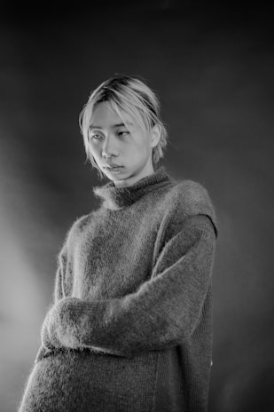 A monochrome portrait of a person with light-colored hair wearing a textured, oversized sweater. The background is blurred, adding focus to the subject's expression, which appears contemplative.