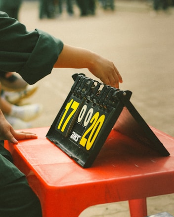 A hand is adjusting the numbers on a scoreboard, which displays the scores 17 and 20. The scoreboard is placed on a red plastic table. The focus is on the hand and the numbers, with a blurred background.