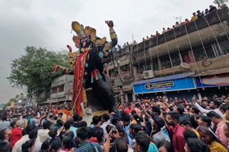 A large crowd gathers on the street for a festival, surrounding a towering statue adorned with elaborate decorations. People on the ground are bustling with excitement, and there are additional onlookers on a building balcony watching the scene. The statue is intricately designed with vibrant colors and detailed embellishments, indicating a cultural celebration.