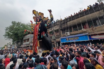 A large crowd gathers on the street for a festival, surrounding a towering statue adorned with elaborate decorations. People on the ground are bustling with excitement, and there are additional onlookers on a building balcony watching the scene. The statue is intricately designed with vibrant colors and detailed embellishments, indicating a cultural celebration.