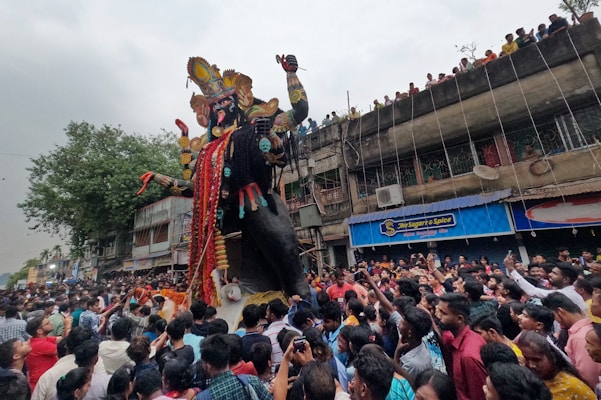 A large crowd gathers on the street for a festival, surrounding a towering statue adorned with elaborate decorations. People on the ground are bustling with excitement, and there are additional onlookers on a building balcony watching the scene. The statue is intricately designed with vibrant colors and detailed embellishments, indicating a cultural celebration.