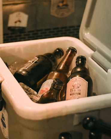 Several beer bottles are nestled in a cooler filled with ice. The cooler is open, revealing a mix of different labeled bottles with condensation visible on their surfaces. The setting has a warm, dimly lit ambiance suggesting a casual or festive atmosphere.