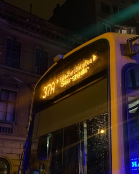 A city bus is seen at night with its LED display showing the route number 37A. The bus is parked in a dimly lit urban environment, visible against the backdrop of old architectural buildings. Streetlights create a warm glow, casting reflections off the wet surfaces of the bus from recent rain.