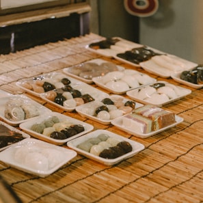 Various trays of traditional Japanese sweets are neatly arranged on a bamboo mat. The confections, likely mochi or dango, exhibit a variety of colors and textures, with some wrapped in plastic. Each tray showcases different assortments, including round and square shapes, with some desserts having a glossy finish.