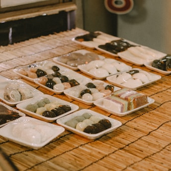 Various trays of traditional Japanese sweets are neatly arranged on a bamboo mat. The confections, likely mochi or dango, exhibit a variety of colors and textures, with some wrapped in plastic. Each tray showcases different assortments, including round and square shapes, with some desserts having a glossy finish.