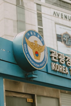 The image features a sign for the Seoul Metropolitan Police Agency, displaying an emblem with a golden eagle and a red circle. The sign is mounted on a blue structure, and there is Korean text prominently visible. In the background, a building with signage that includes 'AVENUE' can be seen. The scene is set in an urban environment.