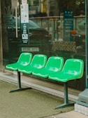 Four green plastic seats are arranged in a row outside a building, possibly near a shop or restaurant. The seats are mounted on metal legs, and the area around them is covered with artificial grass. A glass storefront with signage and menu details is visible in the background.