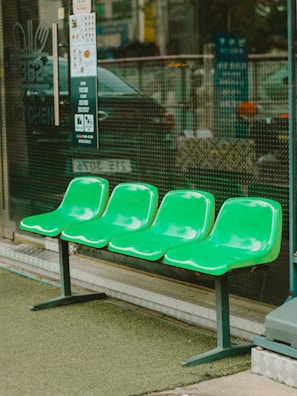 Four green plastic seats are arranged in a row outside a building, possibly near a shop or restaurant. The seats are mounted on metal legs, and the area around them is covered with artificial grass. A glass storefront with signage and menu details is visible in the background.