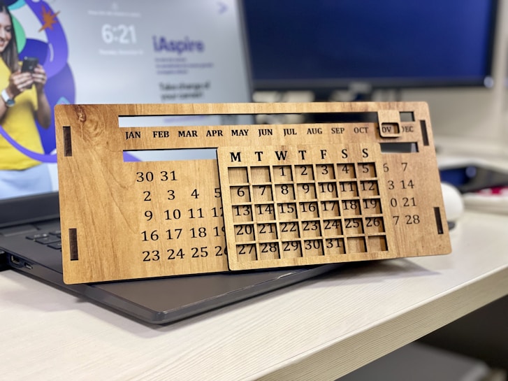 A wooden perpetual calendar with movable parts rests on a dark laptop. It features all the months on the top row and days of the week below, with numbers arranged to represent dates. In the background, a computer monitor displays a blurred digital image of a person holding a phone, with some text and graphics visible.