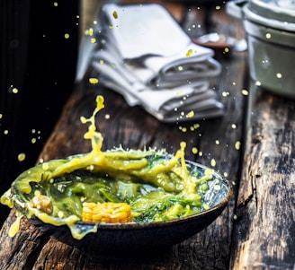 A vibrant splash of green and yellow soup or sauce captured mid-air, with droplets artistically scattering around. A piece of corn is visible in the liquid, resting in a dark, rustic bowl that contrasts with the rich, wooden surface beneath it. In the background, folded white napkins and blurred kitchenware add to the dynamic yet organized composition.