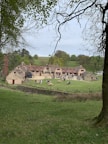 A rustic building with a courtyard is surrounded by lush green fields and trees. Several people are seated at wooden picnic tables enjoying the open space. The atmosphere is peaceful and pastoral.