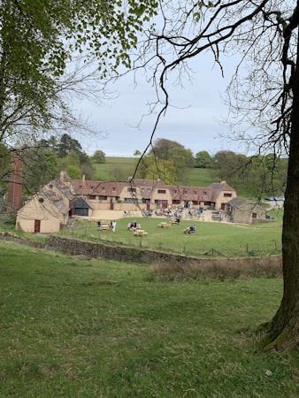 A rustic building with a courtyard is surrounded by lush green fields and trees. Several people are seated at wooden picnic tables enjoying the open space. The atmosphere is peaceful and pastoral.