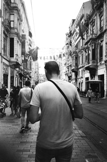 A black and white photograph capturing the back of a man wearing a white t-shirt and a crossbody bag walking down a busy street. The street is lined with tall, ornate buildings and decorated with hanging flags. Several other people are walking along the street, with some pushing strollers. Streetcar tracks run down the center of the cobblestone path.