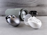 A collection of wireless earbuds is arranged on a flat surface, accompanied by a cylindrical portable speaker in the background. The earbuds vary in color, with one pair being metallic silver and another white, both in stylish, rounded charging cases. The speaker has a fabric exterior with a sleek design.