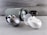 A collection of wireless earbuds is arranged on a flat surface, accompanied by a cylindrical portable speaker in the background. The earbuds vary in color, with one pair being metallic silver and another white, both in stylish, rounded charging cases. The speaker has a fabric exterior with a sleek design.