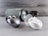 A collection of wireless earbuds is arranged on a flat surface, accompanied by a cylindrical portable speaker in the background. The earbuds vary in color, with one pair being metallic silver and another white, both in stylish, rounded charging cases. The speaker has a fabric exterior with a sleek design.
